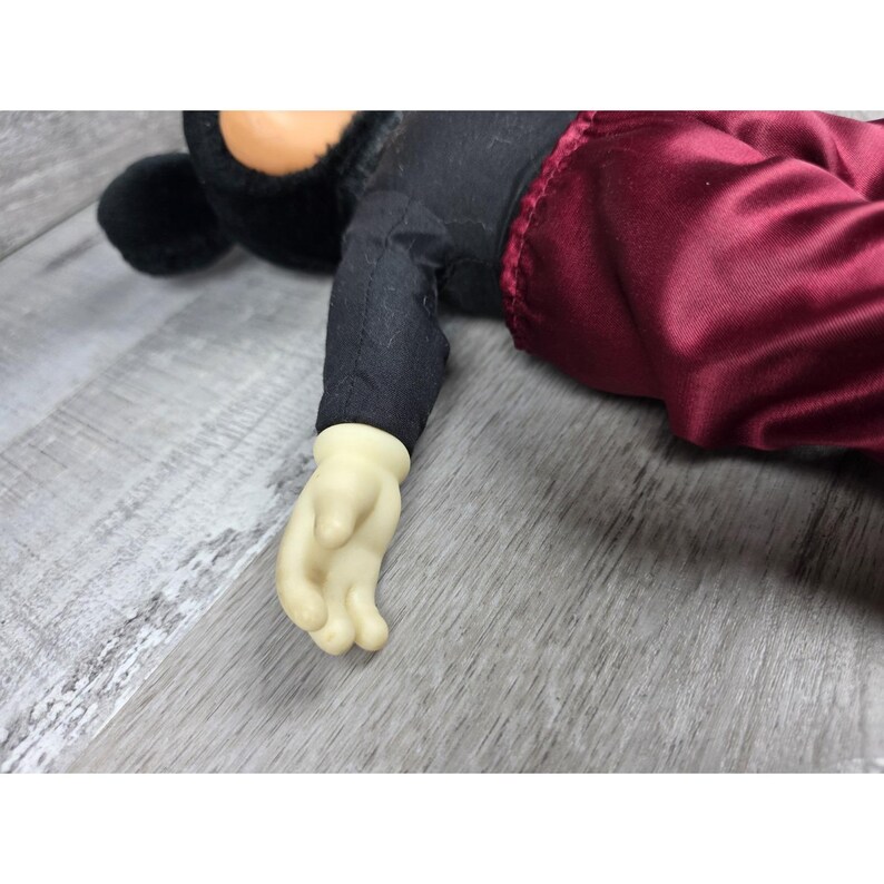 May include: A Mickey Mouse plush toy, featuring a black jacket, red pants, and a cream-colored hand. The plush has a black head and ears. The toy is lying on a gray surface.