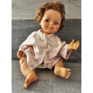 Könnte beinhalten: Eine Vintage-Puppe mit braunen Locken, die ein rosa Kleid mit Blumenmuster und eine Windel trägt. Die Puppe hat ein lächelndes Gesicht mit blauen Augen und rosigen Wangen.