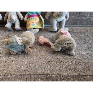 Puede incluir: Una colecci&oacute;n de figuras de animales Epoch Calico Critters vintage. Las figuras incluyen conejos y osos, algunos con ropa. Las figuras est&aacute;n en varias poses, con una combinaci&oacute;n de colores gris claro y azul. La imagen est&aacute; tomada sobre una superficie de madera.