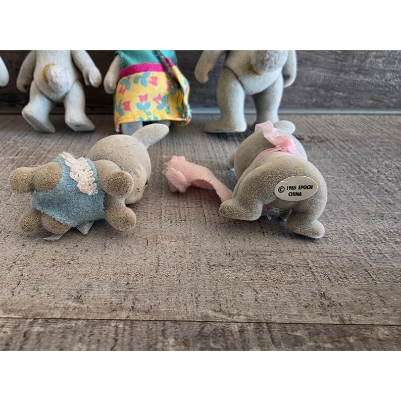 Puede incluir: Una colecci&oacute;n de figuras de animales Epoch Calico Critters vintage. Las figuras incluyen conejos y osos, algunos con ropa. Las figuras est&aacute;n en varias poses, con una combinaci&oacute;n de colores gris claro y azul. La imagen est&aacute; tomada sobre una superficie de madera.