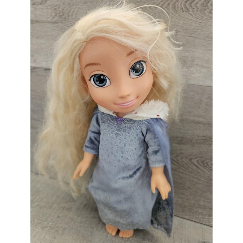 May include: A blonde doll wearing a blue dress with a white fur-trimmed hood. The doll has blue eyes and a smiling face.