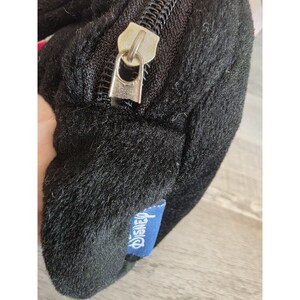 May include: Black plush bag with a silver zipper and a blue Disney tag. The bag has a soft, furry texture. The zipper is open, revealing the interior. The Disney tag is rectangular and has white text.