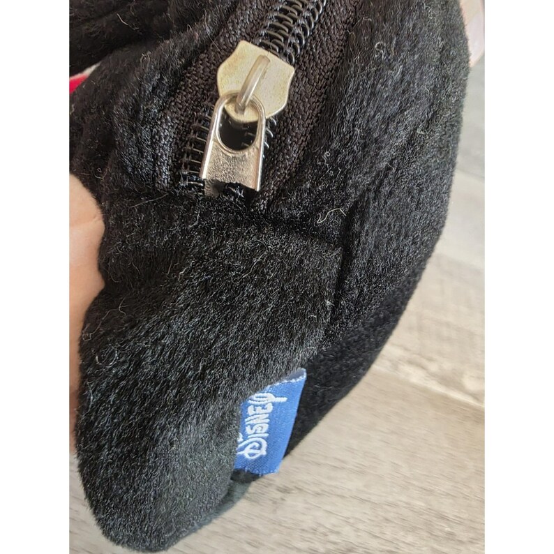 May include: Black plush bag with a silver zipper and a blue Disney tag. The bag has a soft, furry texture. The zipper is open, revealing the interior. The Disney tag is rectangular and has white text.
