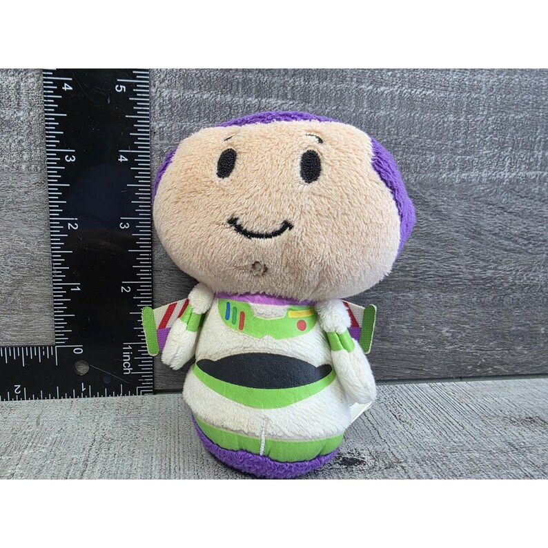 May include: A plush Buzz Lightyear doll, featuring a beige face with a purple head, black eyes, and a stitched smile. The doll is wearing a white and green spacesuit with black accents and purple trim. The doll is approximately 4 inches tall.