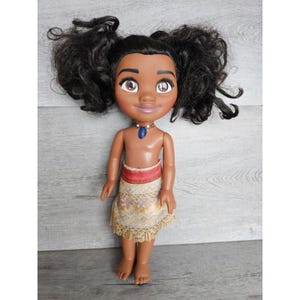 May include: A brown-skinned doll with black curly hair, wearing a brown and gold skirt with a red waistband and a blue necklace. The doll has brown eyes and a smiling face.
