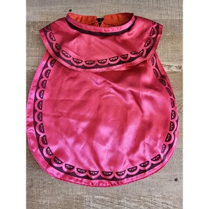 Peut inclure: Une cape en satin rouge avec une bordure noire et un motif noir et blanc. La cape a un col à volants.