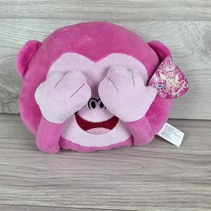 May include: A plush monkey toy in bright pink with a lighter pink face, hands covering its eyes, and a wide smiling mouth. A heart-shaped tag attached to the side displays the text Sugar Loaf Toys with a floral graphic and pink sparkles.