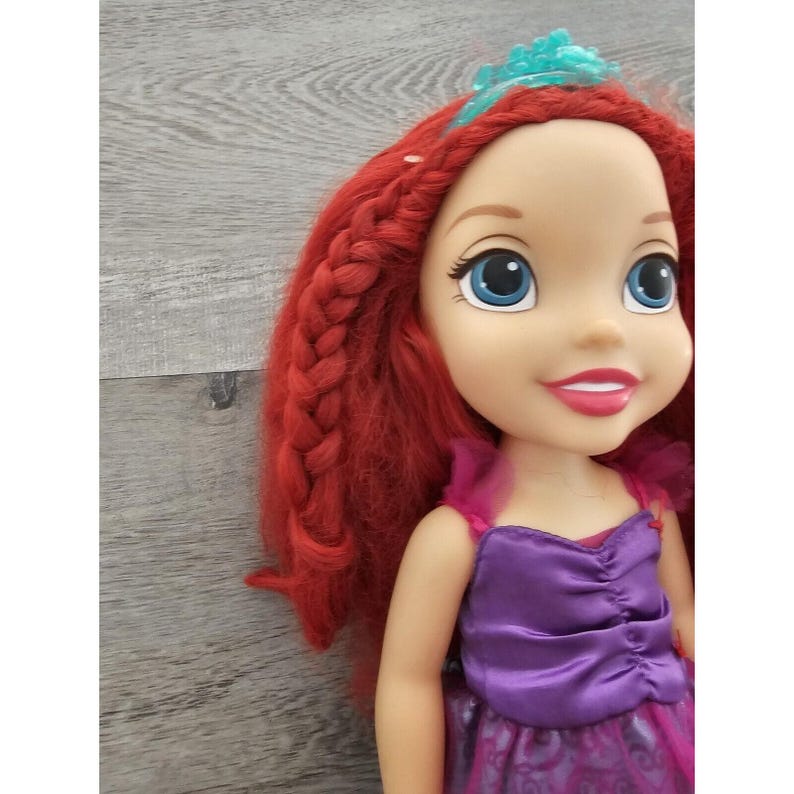 Puede incluir: Una mu&ntilde;eca de pelo rojo con una corona azul, que lleva un vestido morado con estampado floral.