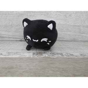 May include: A black plush cat toy with white embroidered eyes and a smiling mouth. The cat is round and has a small, curled tail.