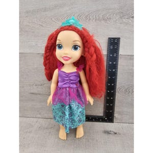 Puede incluir: Una mu&ntilde;eca de pelo rojo con un vestido morado y turquesa con una corona. La mu&ntilde;eca mide aproximadamente 28 cm de alto.