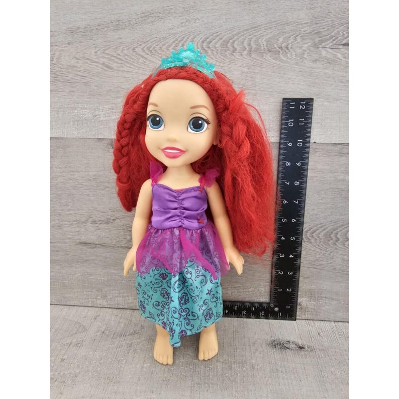 Puede incluir: Una mu&ntilde;eca de pelo rojo con un vestido morado y turquesa con una corona. La mu&ntilde;eca mide aproximadamente 28 cm de alto.