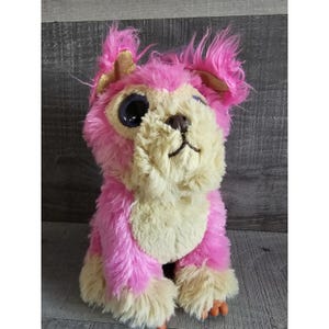 May include: A soft toy dog with pink fur, a cream-coloured face and paws, and large blue eyes. The toy has gold accents on its ears and a small, brown nose. The dog is sitting upright.