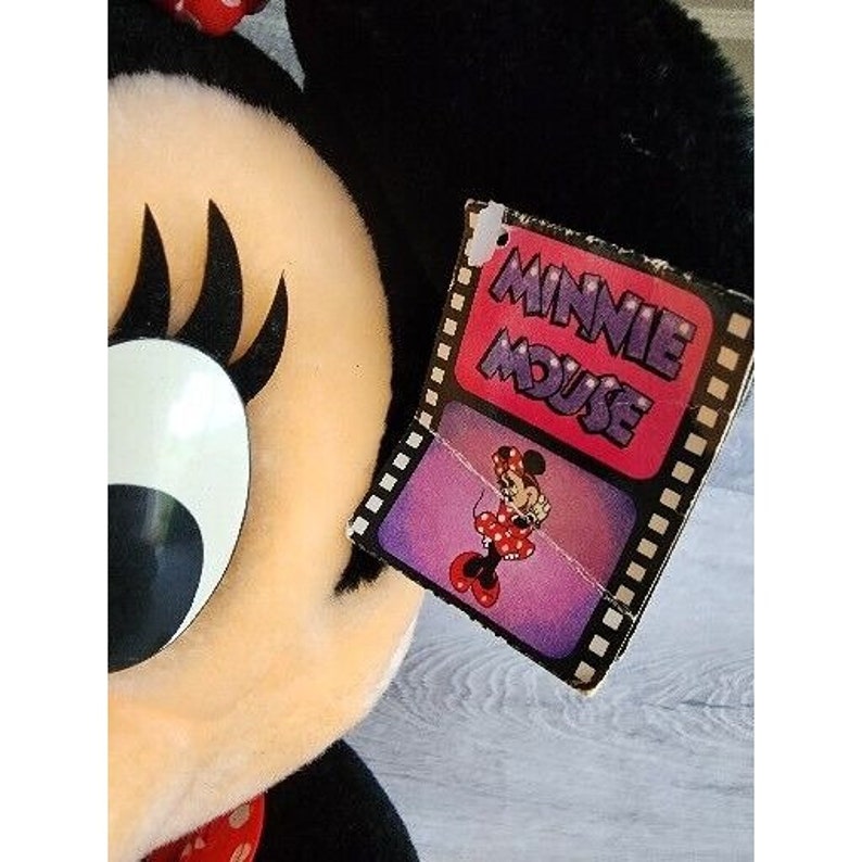 Pu&ograve; includere: Un giocattolo di peluche Minnie Mouse con occhi grandi e un fiocco nero. Il giocattolo ha un'etichetta che recita "Minnie Mouse" con un'immagine di cartone animato di Minnie Mouse.