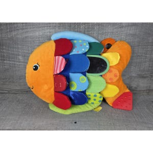 Puede incluir: Un juguete de peluche con forma de pez colorido con una variedad de texturas y patrones. El pez presenta secciones naranjas, azules, verdes y rojas, una cara sonriente y una cola texturizada. El juguete está diseñado para bebés y niños pequeños.