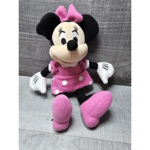 May include: A Minnie Mouse plush toy. The plush has black ears, a pink bow, and a pink dress with white polka dots. The toy has white gloves and pink shoes. The plush is sitting on a gray surface.