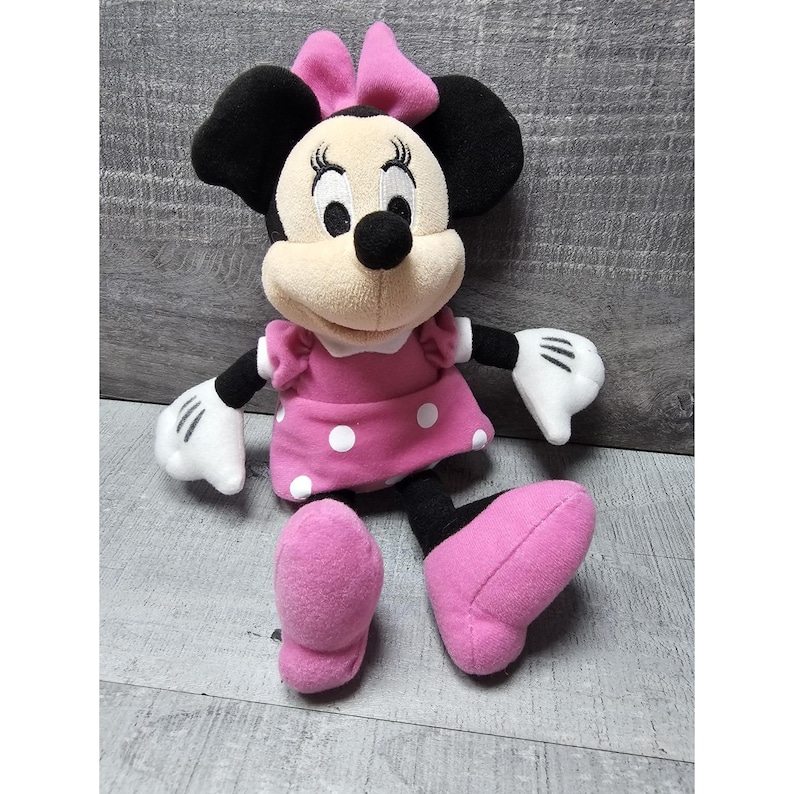 May include: A Minnie Mouse plush toy. The plush has black ears, a pink bow, and a pink dress with white polka dots. The toy has white gloves and pink shoes. The plush is sitting on a gray surface.