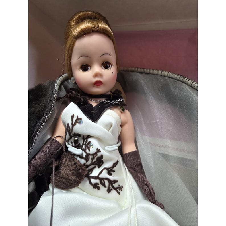 May include: A vintage doll with blonde hair in an updo. The doll is dressed in a white gown with brown floral embroidery and brown gloves. A faux fur wrap and a black choker necklace complete the look. The doll is in a display box.