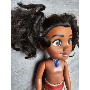 May include: A brown-skinned doll with long, curly black hair styled in two ponytails. The doll is wearing a brown and red skirt and a necklace with a blue gem pendant.