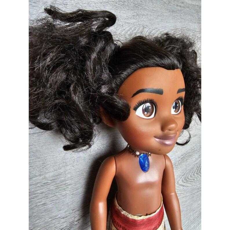 May include: A brown-skinned doll with long, curly black hair styled in two ponytails. The doll is wearing a brown and red skirt and a necklace with a blue gem pendant.