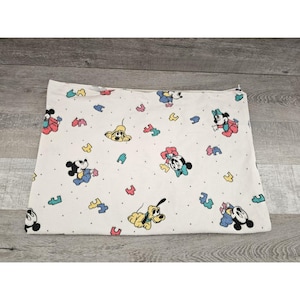 Puede incluir: Una manta de bebé blanca con un patrón repetido de Mickey Mouse, Minnie Mouse y Pluto. Los personajes son de varios colores, incluyendo rojo, azul, amarillo y verde. La manta está cubierta de pequeños puntos blancos.