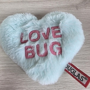 May include: A heart-shaped plush toy made of light mint green faux fur. The words LOVE BUG are embroidered in dark pink thread across the center. A red fabric tag with the white text DOUGLAS is attached to the lower right side of the heart.