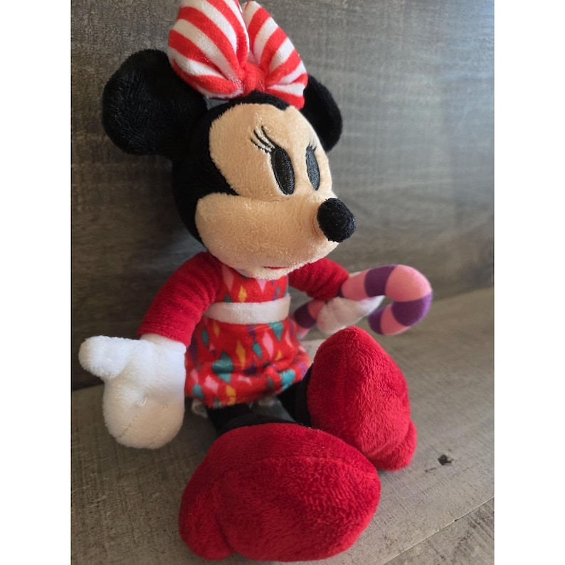 May include: A plush Minnie Mouse doll with a red and white striped bow, black ears, and a red dress with a colorful pattern. She has red shoes and is holding a candy cane. The doll is approximately 10 inches tall.