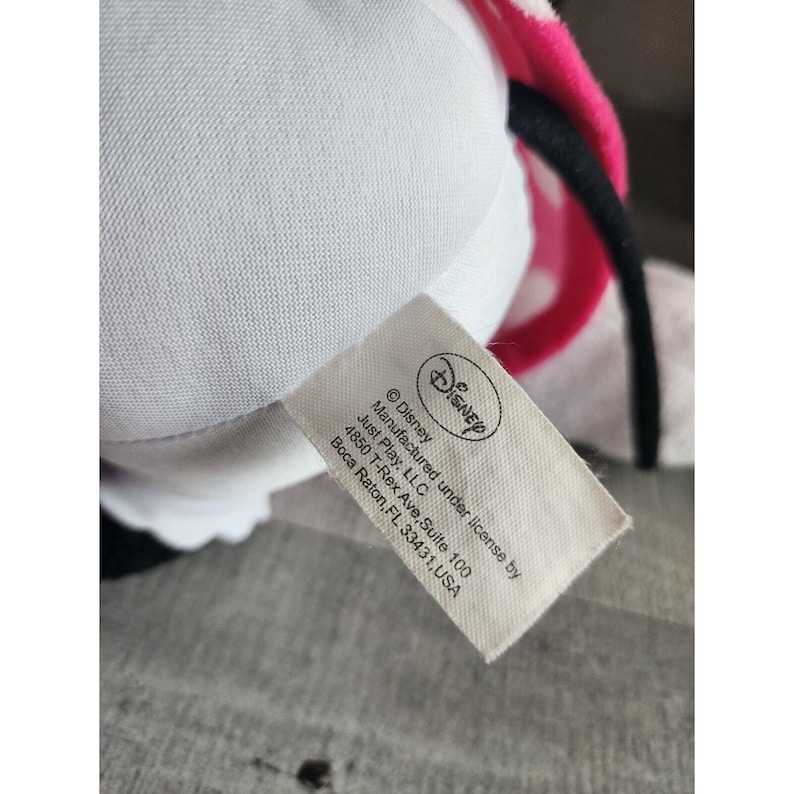 May include: White plush toy with pink and white polka dot fabric and a black bow. The toy has a white tag with the Disney logo and the text "&copy;Disney Manufactured under license by Just Play, LLC 4850 T-Rex Ave, Suite 100 Boca Raton, FL 33431, USA".