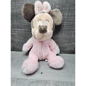 May include: A plush Minnie Mouse doll in a seated position. The doll features a pink body, a light tan face, and large brown ears. The doll has embroidered facial features, including eyelashes and a nose. The doll is made of soft, plush material.