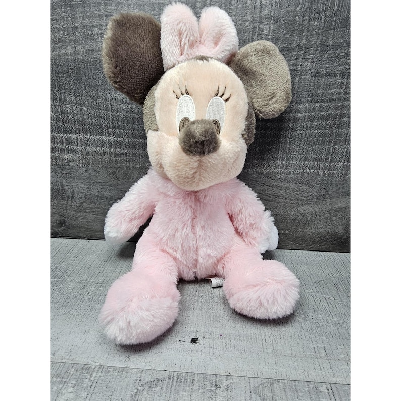 May include: A plush Minnie Mouse doll in a seated position. The doll features a pink body, a light tan face, and large brown ears. The doll has embroidered facial features, including eyelashes and a nose. The doll is made of soft, plush material.