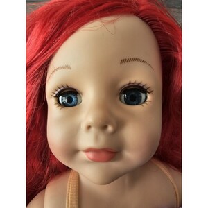 Puede incluir: Primer plano de la cara de una mu&ntilde;eca con cabello rojo, ojos azules y boca rosa claro. La mu&ntilde;eca lleva un top marr&oacute;n claro sin mangas.