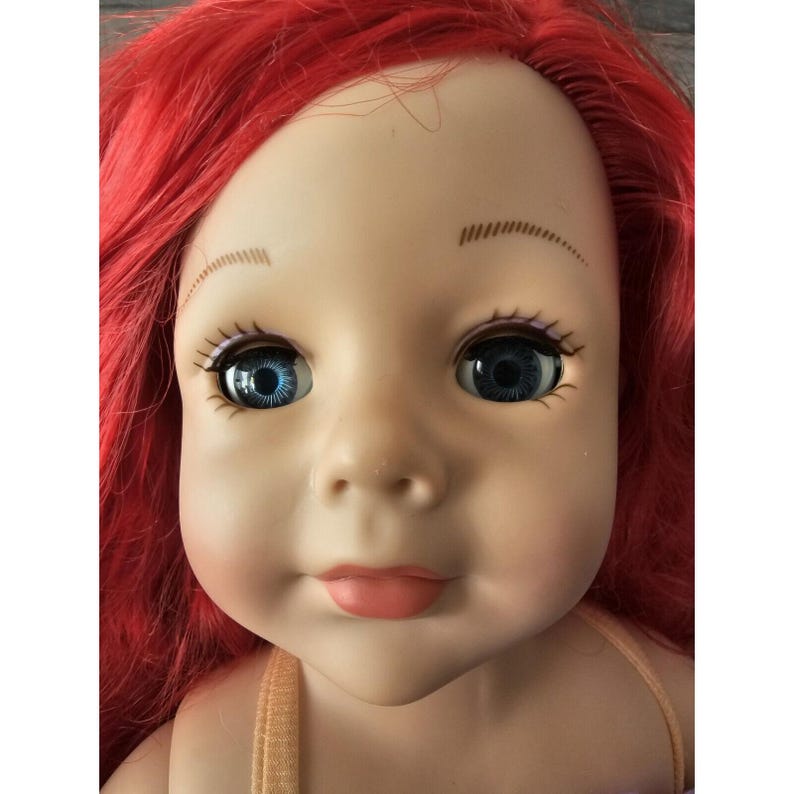 Puede incluir: Primer plano de la cara de una mu&ntilde;eca con cabello rojo, ojos azules y boca rosa claro. La mu&ntilde;eca lleva un top marr&oacute;n claro sin mangas.