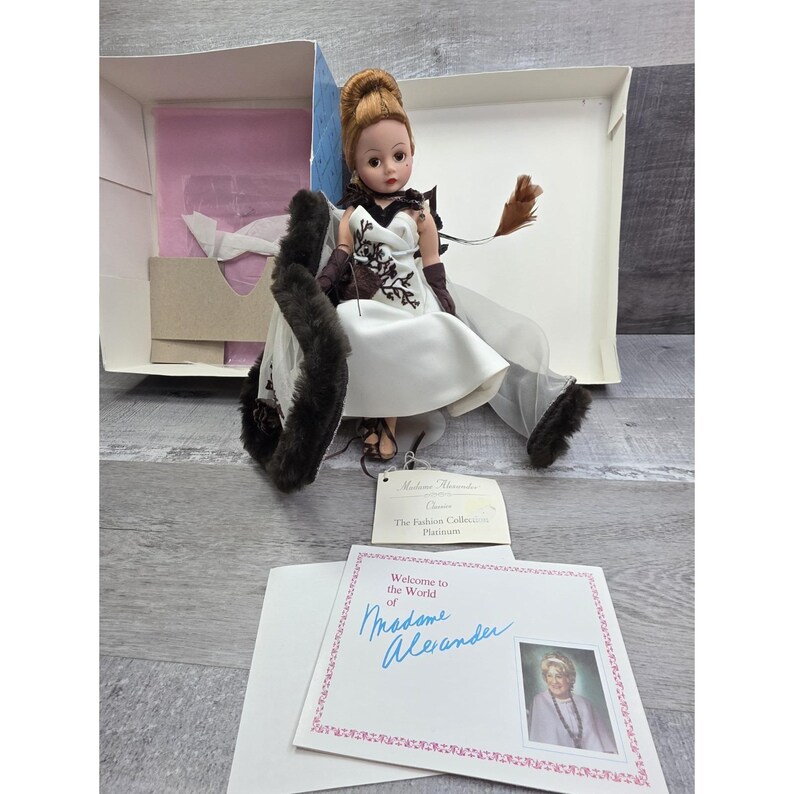 May include: A Madame Alexander doll from The Fashion Collection Platinum series. The doll is dressed in a white satin dress with a black and brown floral design, brown gloves, and a fur stole. The doll is in its original box with a welcome card.