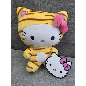 May include: A plush Hello Kitty doll dressed in a tiger costume. The doll is yellow with brown stripes, a white face, and a pink bow. A white tag with a Hello Kitty graphic and pink bow is attached.