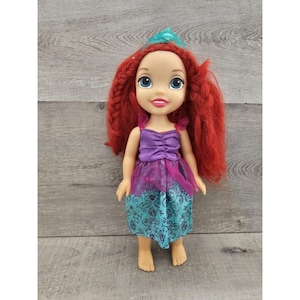 Puede incluir: Una mu&ntilde;eca de pelo rojo con un vestido morado y turquesa con una corona. La mu&ntilde;eca tiene una cara sonriente con ojos azules.