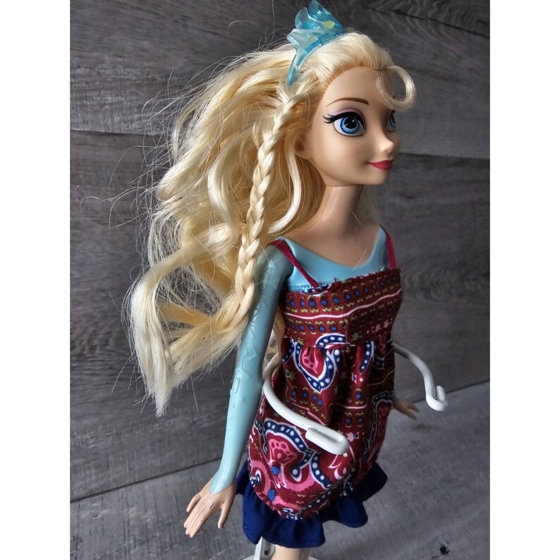 Puede incluir: Una mu&ntilde;eca rubia con ojos azules que lleva un vestido estampado en rojo y azul y una corona azul.
