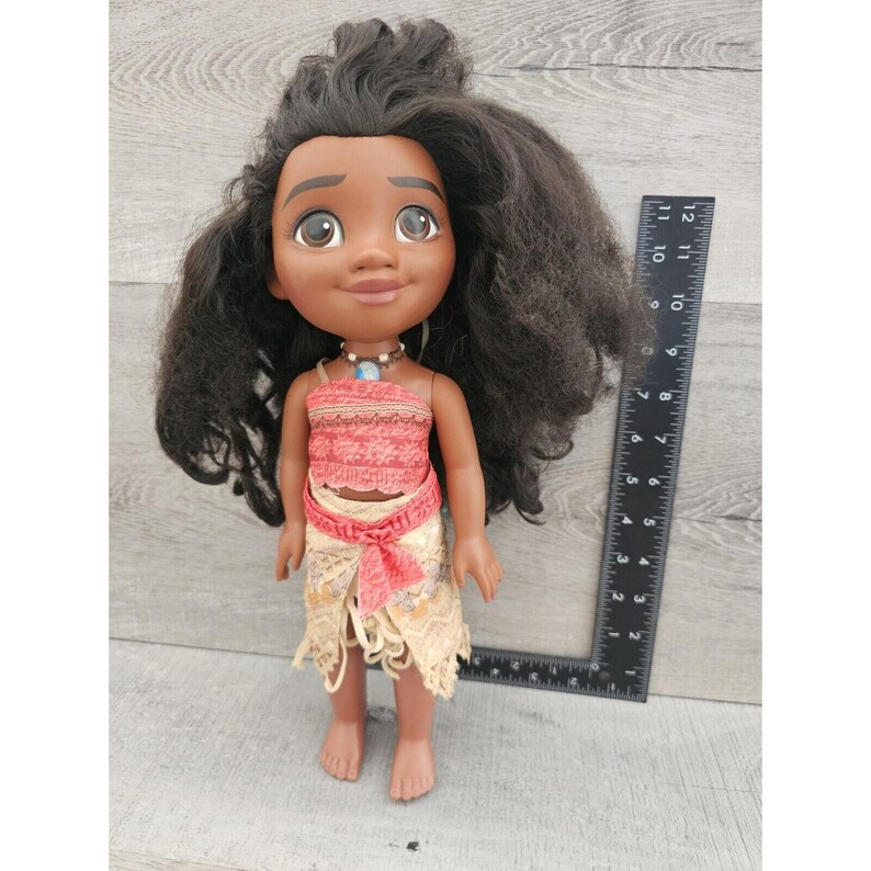 Puede incluir: Una mu&ntilde;eca de piel morena con el pelo largo y negro rizado que lleva un top rojo y blanco y una falda beige con un lazo rojo. La mu&ntilde;eca mide aproximadamente 28 cm de altura.