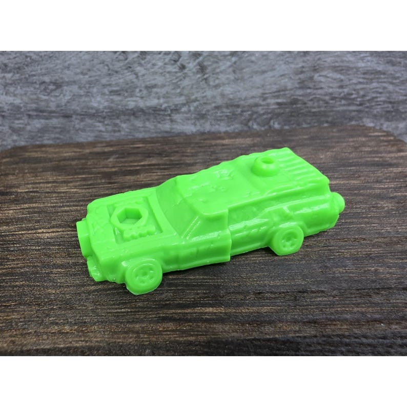 以下が含まれることがあります： 鮮やかな緑色の玩具の車。四角いデザインで、車輪と上部に2つの円形の開口部があります。おもちゃの車はプラスチック製で、濃い木製の表面に置かれています。