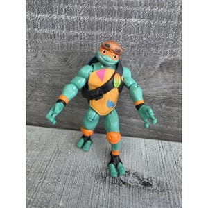 May include: A Teenage Mutant Ninja Turtle action figure. The figure is green with orange accents, a brown helmet, and a yellow and orange chest plate. The figure is standing on a grey surface.