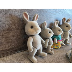 Puede incluir: Una colecci&oacute;n de figuras de conejos grises con orejas largas y pelaje suave. Un conejo lleva un vestido turquesa y amarillo con estampado floral. Los conejos est&aacute;n de pie, con los brazos a los lados, mirando hacia la derecha.