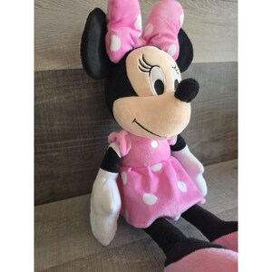 May include: A plush Minnie Mouse doll with a pink dress and bow, both adorned with white polka dots. The doll has black ears, arms, and legs, and white gloves. The doll is sitting and smiling.