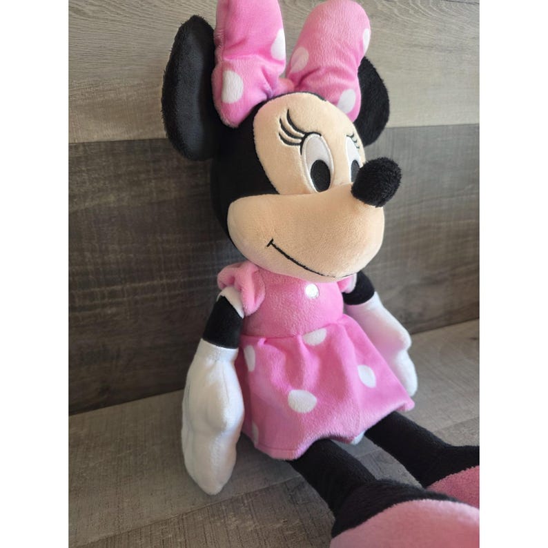 May include: A plush Minnie Mouse doll with a pink dress and bow, both adorned with white polka dots. The doll has black ears, arms, and legs, and white gloves. The doll is sitting and smiling.