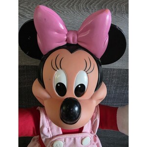 May include: A Minnie Mouse plush doll with a pink bow, black ears, and a pink and red outfit. The doll has large, expressive eyes and a round, black nose. The doll's outfit includes a pink dress with white buttons.