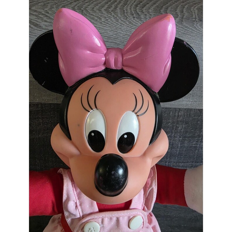 May include: A Minnie Mouse plush doll with a pink bow, black ears, and a pink and red outfit. The doll has large, expressive eyes and a round, black nose. The doll's outfit includes a pink dress with white buttons.