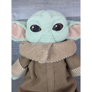 May include: A plush doll of a character with large eyes, large ears, and a tan coat. The doll has a light green face and ears with pink inner ears. The doll's coat is brown with a corduroy collar.