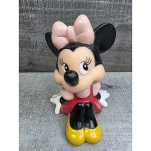 May include: A Minnie Mouse figurine with a pink bow, black ears, and a red dress. The figurine has yellow shoes and white gloves. The figure is sitting down and has a cheerful expression.