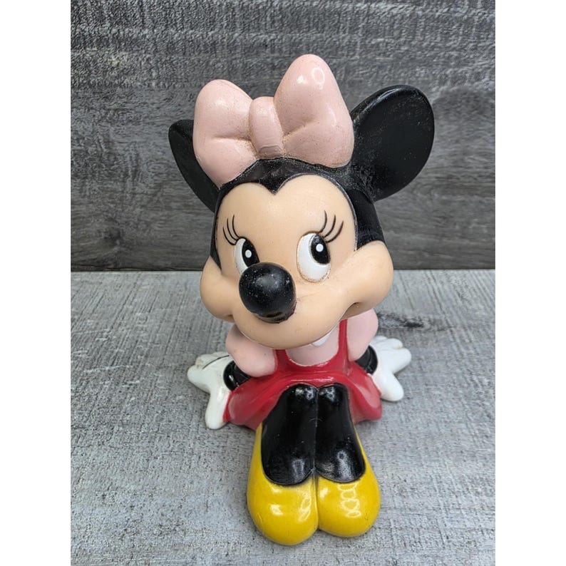 May include: A Minnie Mouse figurine with a pink bow, black ears, and a red dress. The figurine has yellow shoes and white gloves. The figure is sitting down and has a cheerful expression.