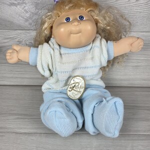 May include: A doll with blonde hair, blue eyes, and a purple hair accessory. The doll wears a white and light blue striped knit sweater and matching light blue knit pants. A gold-toned tag with the text Renzo and a map of Italy is attached.