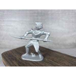 May include: A silver-colored toy knight figurine, posed with a spear. The knight is wearing detailed armor and a helmet. The figurine is standing on a dark wooden surface against a gray background.