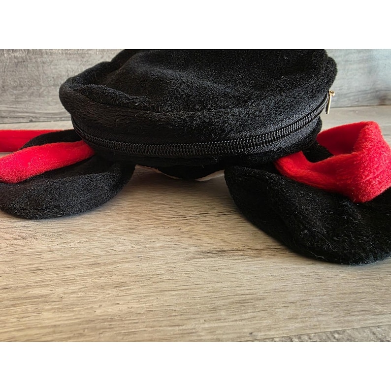 May include: Black plush bag with a zippered closure and two attached black and red plush elements. The bag is on a wooden surface.