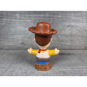 May include: A small plastic toy figure of a cowboy character, wearing a brown hat, yellow shirt, blue pants, and a red neckerchief. The figure has outstretched arms and is standing on a gray surface.